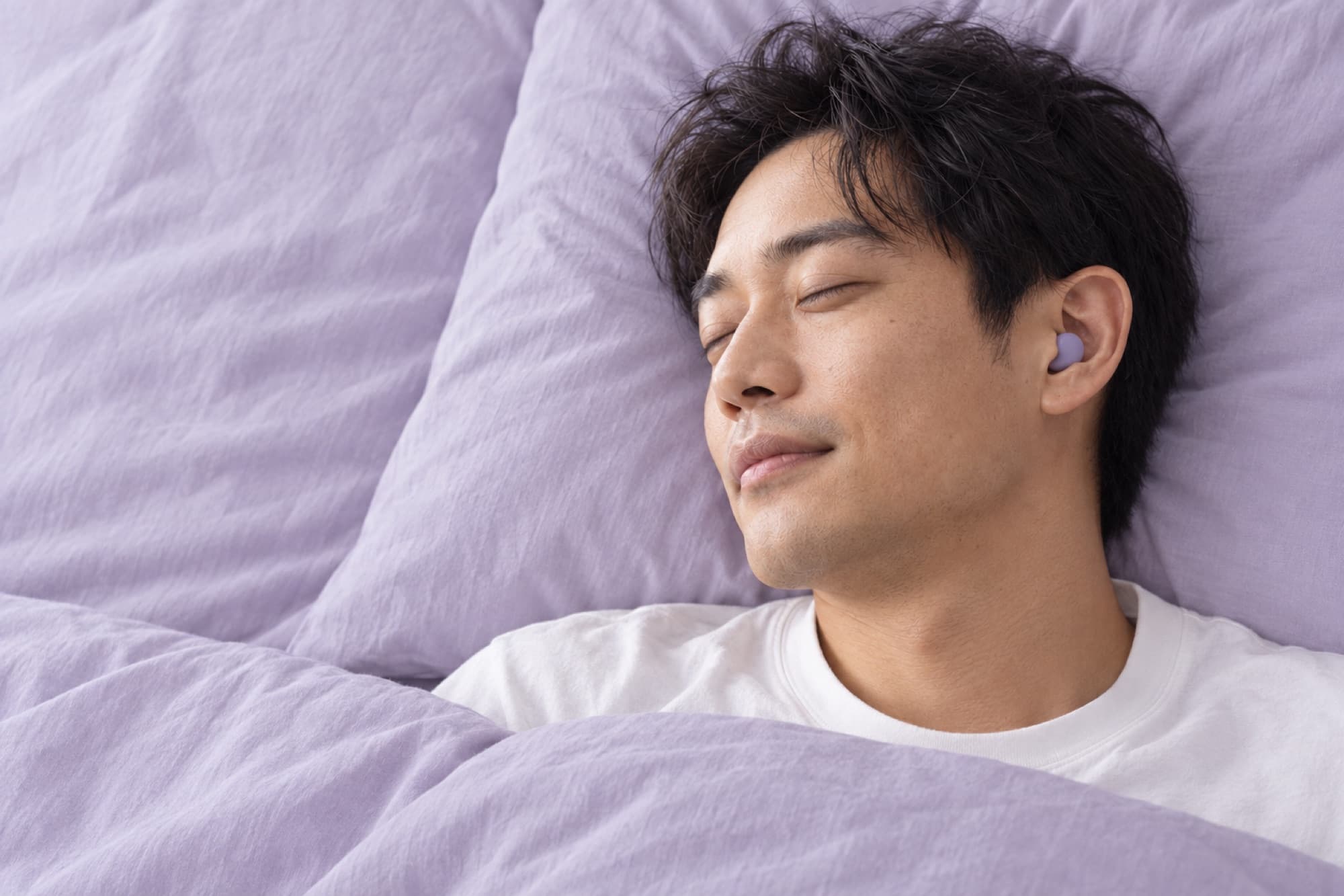 ラベンダー色の寝具で、イヤプラグをつけて目を閉じている男性